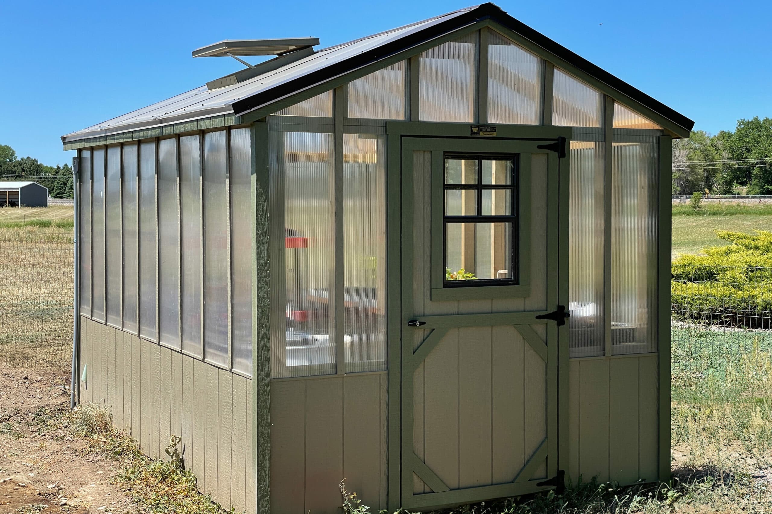 tan and green trimmed clear sided greenhouse shed with plants inside and vent on roof